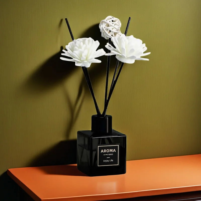 Black aroma diffuser bottle with white artificial flowers and reed sticks on orange table