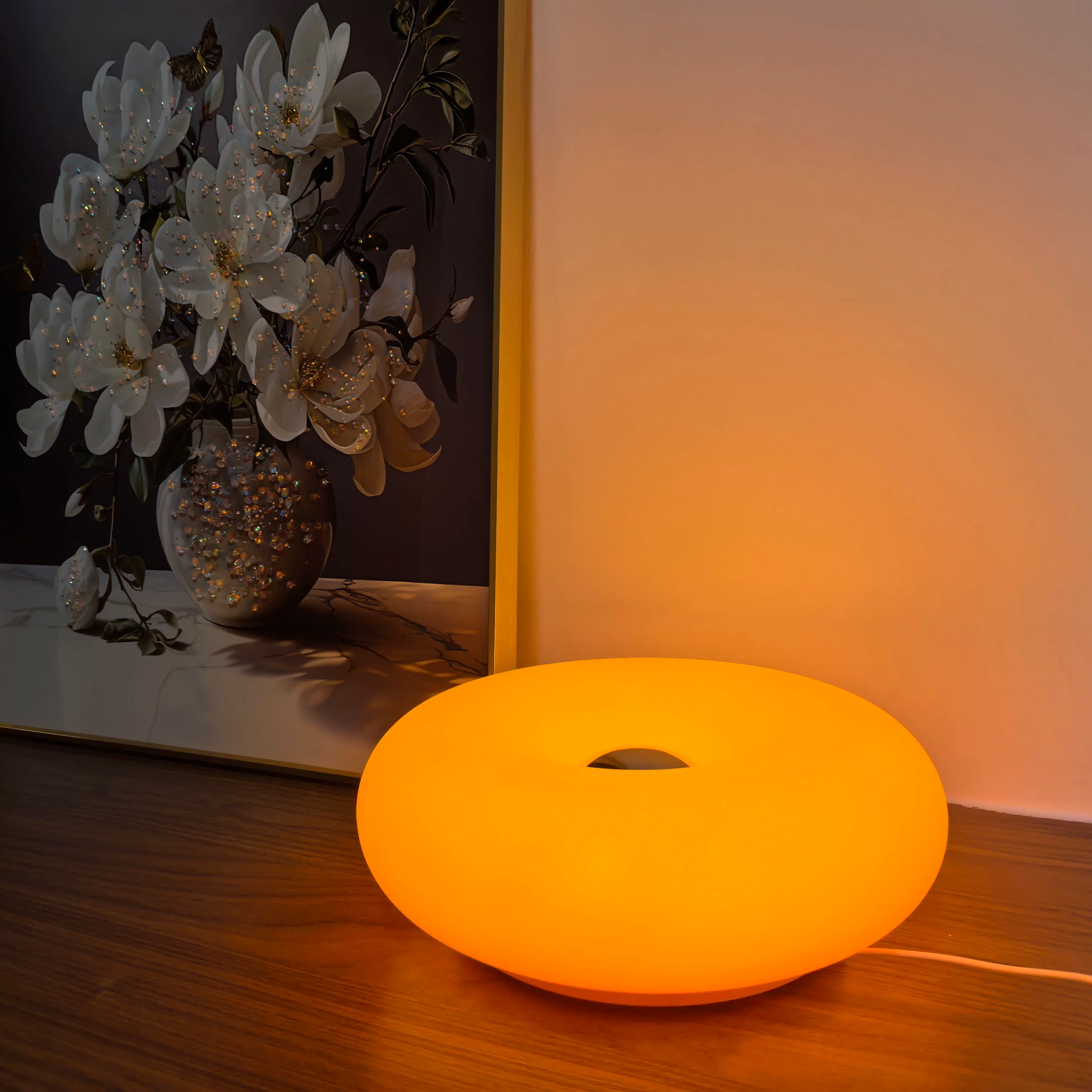 Modern donut-shaped orange lamp on wooden table beside floral painting, ambient lighting