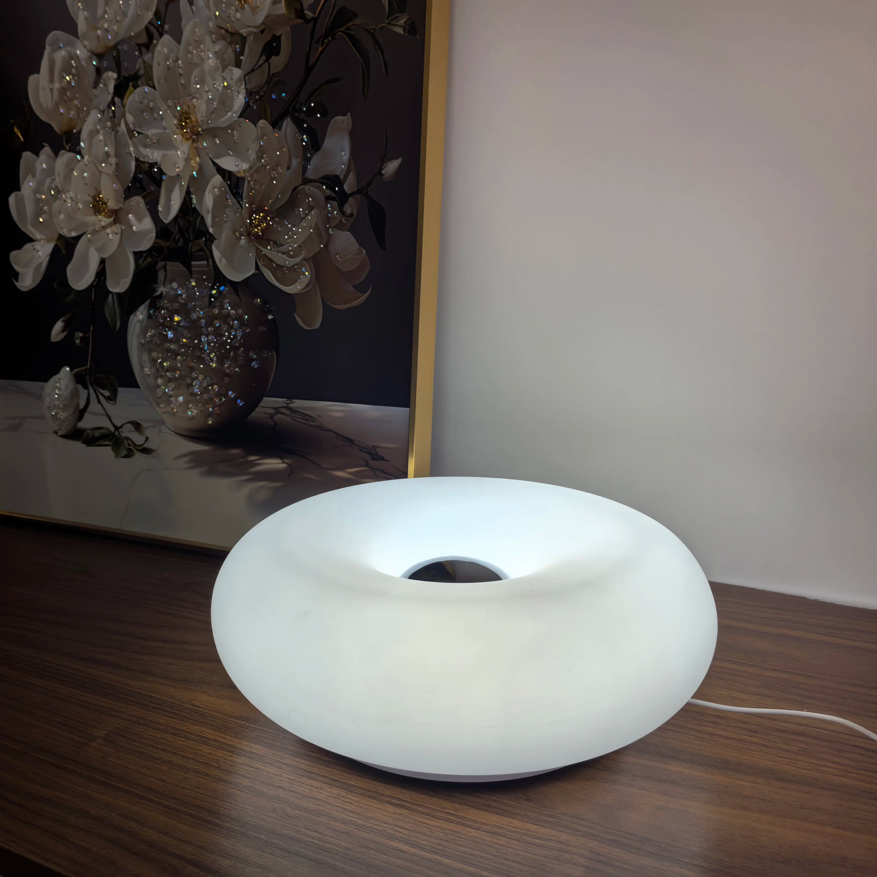 Modern white donut-shaped lamp on wooden table, floral painting in background